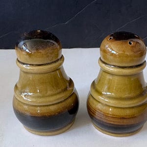 May include: A pair of ceramic salt and pepper shakers. The shakers are cylindrical with a bulbous top and base. The color scheme is a gradient of brown, tan, and black. The salt shaker has three holes on top.