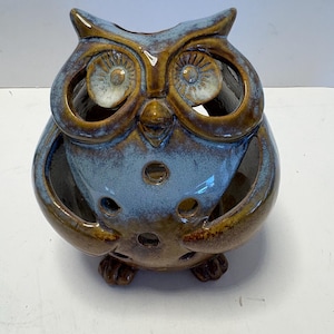 May include: A ceramic owl-shaped candle holder with a blue and brown glaze. The owl has large eye openings, a beak, and small feet. The body features several small circular openings. This decorative item is suitable for home decor.
