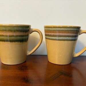 May include: Two ceramic mugs with brown, green, and pink striped designs. The mugs have a light brown glaze and are perfect for coffee or tea.