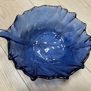 Op de afbeelding: Een diepblauwe glazen kom in de vorm van een blad, met een textuur en een handvat. De kom heeft een geschulpte rand. Een decoratief item.
