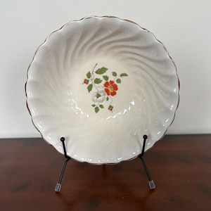 Könnte beinhalten: Eine weiße Keramikschüssel mit goldfarbenem Rand und einem Blumendesign mit roten und weißen Blüten. Die Schüssel steht auf einem schwarzen Ständer.