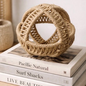 Puede incluir: Una esfera decorativa tejida con cuerda de color natural se encuentra sobre una pila de libros. La esfera tiene un diseño geométrico abierto. Los libros tienen títulos como "Pacific Natural" y "Surf Shacks".