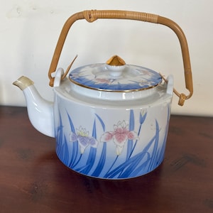 Può includere: Una teiera in porcellana bianca con un motivo floreale blu e bianco. La teiera ha un manico in bambù e un bordo dorato.