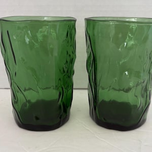 Puede incluir: Dos vasos verdes con un diseño texturizado y orgánico. Los vasos tienen una base verde oscuro y una superficie ondulada e irregular. Los vasos son de vidrio y están vacíos.