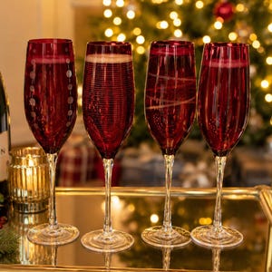 Peut inclure: Quatre flûtes à champagne rouge, remplies d'une boisson pétillante, sont disposées sur un chariot doré. Une bouteille de champagne est visible. En arrière-plan, un sapin de Noël illuminé de lumières scintillantes.
