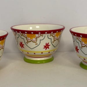 Op de afbeelding: Drie keramische kommen met een witte achtergrond, rode randen en groene bodems. De kommen hebben een bloemenontwerp met gele, rode en groene accenten. De kommen zijn geschikt voor het serveren van voedsel.