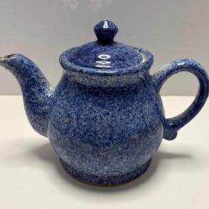 May include: A blue ceramic teapot with a speckled pattern. The teapot has a lid with a small knob, a curved spout, and a rounded handle. The teapot is designed for brewing tea.