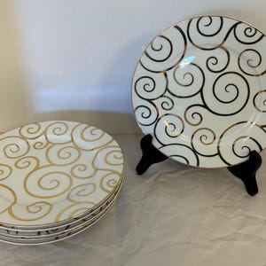 May include: A set of white ceramic plates featuring a repeating pattern of metallic gold swirls. One plate is displayed upright on a black plastic stand, while three additional plates are stacked horizontally in a pile to the left side.