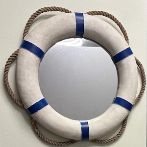 May include: A round mirror framed by a cream-colored life preserver with blue stripes. A thick, brown rope encircles the life preserver, adding a nautical touch. The mirror reflects the surrounding environment.