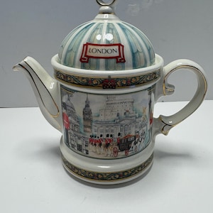 Könnte beinhalten: Dekorative Keramik-Teekanne mit London-Motiv. Der Deckel ist blau-weiß gestreift mit dem Wort "LONDON" auf einem roten Banner. Der Körper der Teekanne zeigt ikonische Londoner Wahrzeichen und Figuren, mit goldenen Zierleisten.