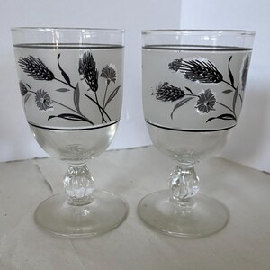 May include: Two clear glass goblets featuring a wide, frosted gray band around the upper portion. The band is decorated with a black and white botanical print of wheat stalks and small flowers, accented by thin black horizontal lines.