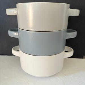 May include: A stack of three ceramic soup bowls in shades of white, gray, and light gray. Each bowl has two small handles. The bowls are stacked on top of each other, with the white bowl at the bottom.