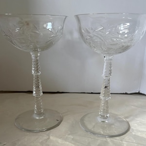 Könnte beinhalten: Zwei klare Glasschalen mit geätzten Blumenmustern auf den Kuppen und gestuften, strukturierten Stielen. Jedes Glas hat eine breite, flache Schale und einen runden Fuß, wobei die Stielabschnitte ein detailliertes, facettiertes Design aufweisen.