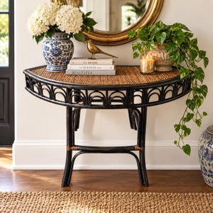 May include: A semi-circular black rattan console table with a woven top. On the table are a blue and white ceramic vase with white hydrangeas, a gold bird figurine, three stacked books titled At Home, The Kinfolk Home, and Beautiful, and a candle.