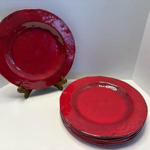 Könnte beinhalten: Ein Set roter Keramikplatten mit einem Used-Look-Finish. Eine Platte wird auf einem Holzständer präsentiert, die anderen sind gestapelt. Die Platten haben einen leicht unregelmäßigen Rand und eine glänzende Oberfläche.