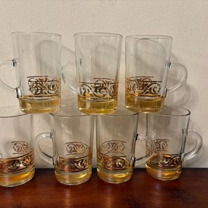 May include: Set of seven clear glass mugs with handles, each featuring a gold decorative band near the base. The mugs are filled with a golden liquid, creating a warm, inviting aesthetic. Ideal for serving beverages.