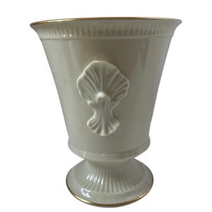 Peut inclure: Un vase en céramique crème avec un motif de coquillage. Le vase a un dessus évasé avec un bord doré et une base en piédestal. Le coquillage est centré sur le devant. Un objet décoratif.
