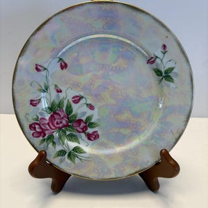 Pode incluir: Um prato decorativo com acabamento perolado e borda dourada. O prato apresenta um desenho floral de rosas cor-de-rosa e folhas verdes. O prato é exibido em um suporte de madeira.