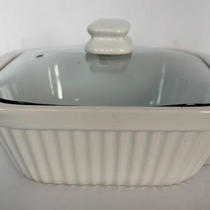 Peut inclure: Un plat à gratin rectangulaire blanc avec un couvercle en verre. Le plat présente un motif côtelé sur les côtés et un bouton blanc sur le couvercle. Le couvercle en verre reflète la lumière.