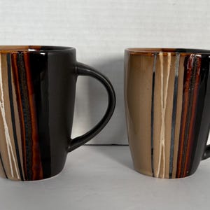 May include: Two ceramic mugs with a unique design. One mug is primarily black, the other is tan. Both mugs have a curved handle and vertical stripes in shades of brown, tan, and white. The mugs are suitable for coffee, tea, or other beverages.