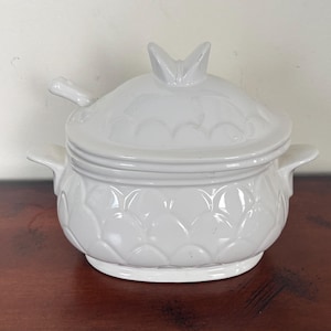 May include: A white ceramic serving dish with a lid and a small spoon. The dish has a textured, scalloped design on the body and a decorative handle on the lid. The dish is sitting on a brown surface.