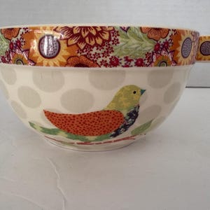 Puede incluir: Un cuenco de sopa de cerámica con asa, con una base crema con grandes lunares grises. El cuenco está decorado con un colorido patrón floral y de pájaros alrededor del borde. El pájaro tiene detalles naranjas, verdes y azules.