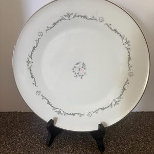 May include: A round white ceramic dinner plate featuring a silver rim and a delicate gray scrollwork border pattern. The center of the plate displays a small, detailed cluster of pink and white roses with green leaves, creating a balanced design.