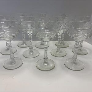 Könnte beinhalten: Eine Sammlung klarer Champagnergläser. Jedes Glas hat eine breite, flache Schale und einen dekorativen, strukturierten Stiel. Die Gläser sind auf einer weißen Oberfläche angeordnet, was ihr elegantes Design zeigt. Ideal für besondere Anlässe.