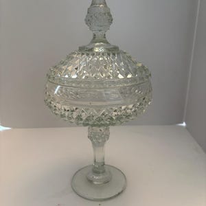 May include: A clear, vintage-style glass candy dish with a lid. The dish features a diamond-cut pattern and sits on a pedestal base. The lid has a pointed finial. This decorative piece is suitable for storing small items.
