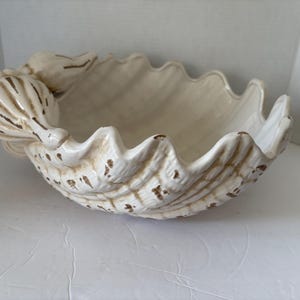 May include: Cream-colored ceramic seashell bowl with a textured surface and scalloped edges. The bowl has brown accents and a detailed seashell design, perfect for coastal decor or serving dishes.