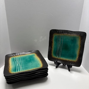 Może przedstawiać: Zestaw sześciu kwadratowych talerzy ceramicznych. Każdy talerz ma czarną obwódkę ze złotym wykończeniem, otaczającą turkusowy środek. Białe pionowe linie zdobią turkusową powierzchnię. Jeden talerz stoi na czarnym stojaku.