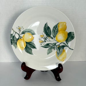 Puede incluir: Plato de cerámica blanca con diseño de rama de limón. El plato presenta limones amarillos, hojas verdes y flores blancas. Se exhibe sobre un soporte de madera marrón oscuro, mostrando un diseño clásico con temática de frutas.