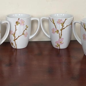 May include: Six white ceramic mugs with a floral design. Each mug features a brown branch with pink blossoms. The mugs have a curved shape and a large handle. The mugs are displayed on a dark brown surface.