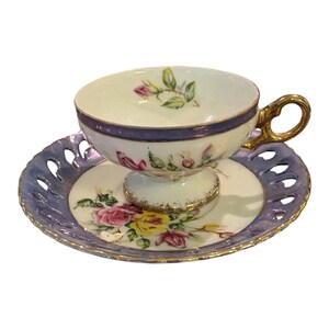 May include: A porcelain teacup and saucer set featuring a white base with hand-painted pink and yellow roses. The saucer has a purple iridescent border with a decorative lattice cutout edge. The teacup includes a gold handle and purple trim.