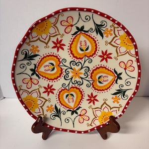 May include: A decorative ceramic plate with a cream-colored background and a vibrant floral design. The plate features a red rim with white polka dots and is adorned with yellow, orange, and red flowers and heart-shaped motifs. The plate is displayed on a wooden stand.