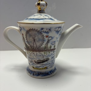 May include: A white ceramic teapot with a lid and a gold crown-shaped handle. The teapot features a blue and white design depicting the London Eye and the Thames River. The base reads "Thames Riverside London".