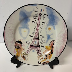 May include: A decorative plate featuring the Eiffel Tower, with the text "La Tour Eiffel". The plate has a black rim and a design with pink, blue, and yellow accents. The plate is supported by a black stand.
