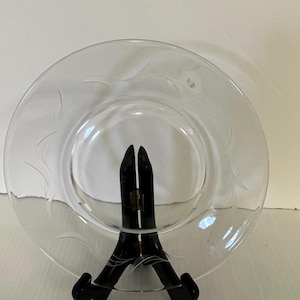 May include: A clear glass plate featuring a delicate etched leaf pattern along the rim. The plate is circular with a smooth, transparent surface and is supported by a black folding display stand that holds it in an upright position.