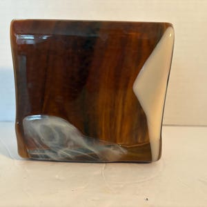 May include: A handcrafted glass art piece, rectangular in shape, featuring a blend of brown and amber hues with a white accent. The glass has a glossy finish and a unique, organic design. This decorative item could be used as a small vase or a decorative object.