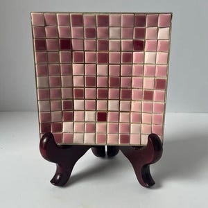 Könnte beinhalten: Ein rechteckiges dekoratives Tablett mit einem Mosaikfliesen-Design in Rosa- und Burgundertönen. Das Tablett wird auf einem dunkelbraunen Holzständer präsentiert. Die Mosaikfliesen sind in einem Rastermuster angeordnet.