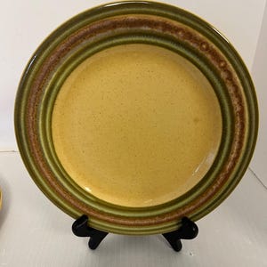 May include: A round ceramic plate featuring a speckled mustard yellow center. The rim is decorated with concentric bands of olive green and a textured, earthy brown ring. The plate has a glossy finish and is displayed on a black stand.