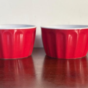 Może przedstawiać: Cztery czerwone ceramiczne kokilki z białym wnętrzem ustawione na ciemnej drewnianej powierzchni. Kokilki mają żebrowany wygląd zewnętrzny. Tło to gładka biała ściana.