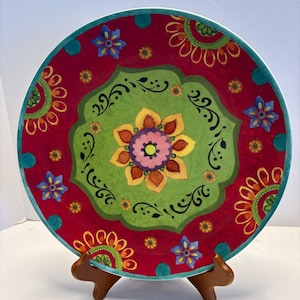 May include: A colourful ceramic plate with a floral design. The plate has a red background with a green central design and a turquoise rim. The floral pattern includes yellow, pink, and blue accents. The plate is displayed on a wooden stand.