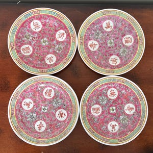 May include: Set of four round ceramic coasters. Each coaster features a pink background with floral and Chinese character designs. The coasters have a white border and a decorative green and yellow outer edge.