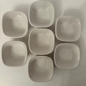 May include: A top-down shot of seven small, off-white ceramic bowls. Each bowl is square with rounded corners and a slightly recessed interior. The bowls are arranged in a circular pattern on a white surface.