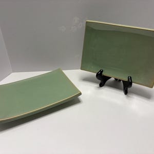 May include: Two rectangular, sage green ceramic plates with a slightly curved design. One plate is on a black stand. The plates have a gold-colored trim. The plates are likely for serving food.
