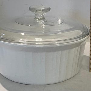 May include: A white, round casserole dish with a clear glass lid and a clear glass knob. The dish has vertical ribbed detailing. A wooden ruler is next to the dish.