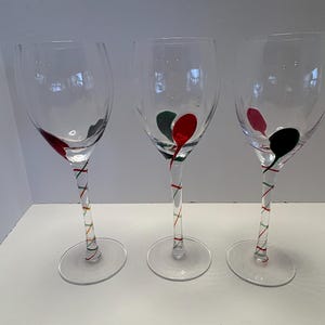 May include: Three clear wine glasses with colorful accents. Each glass has a twisted stem with red, green, and gold stripes. The bowl of each glass features a splash of red, green, and black paint.
