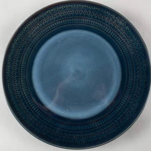 Op de afbeelding: Een ronde, blauwe keramische plaat met een getextureerd oppervlak. De plaat heeft een reliëf geometrisch patroon rond de rand, inclusief stippen, ruiten en lijnen. Het midden van de plaat is effen, donkerder blauw.