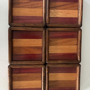 Può includere: Sei sottobicchieri quadrati in legno con una cornice marrone scuro e un design a strisce. Le strisce alternano tra tonalità di legno marrone rossiccio e marrone chiaro. I sottobicchieri sono disposti a griglia.